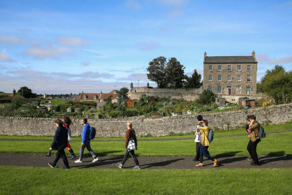 People walk along a path in front of a stone wall. The buildings of Berwick are in the background.