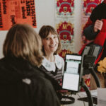 Smiling woman and the back of another persons head sat in front of a touch screen. Posters of the bright pink Berwick bear Festival poster in the background.