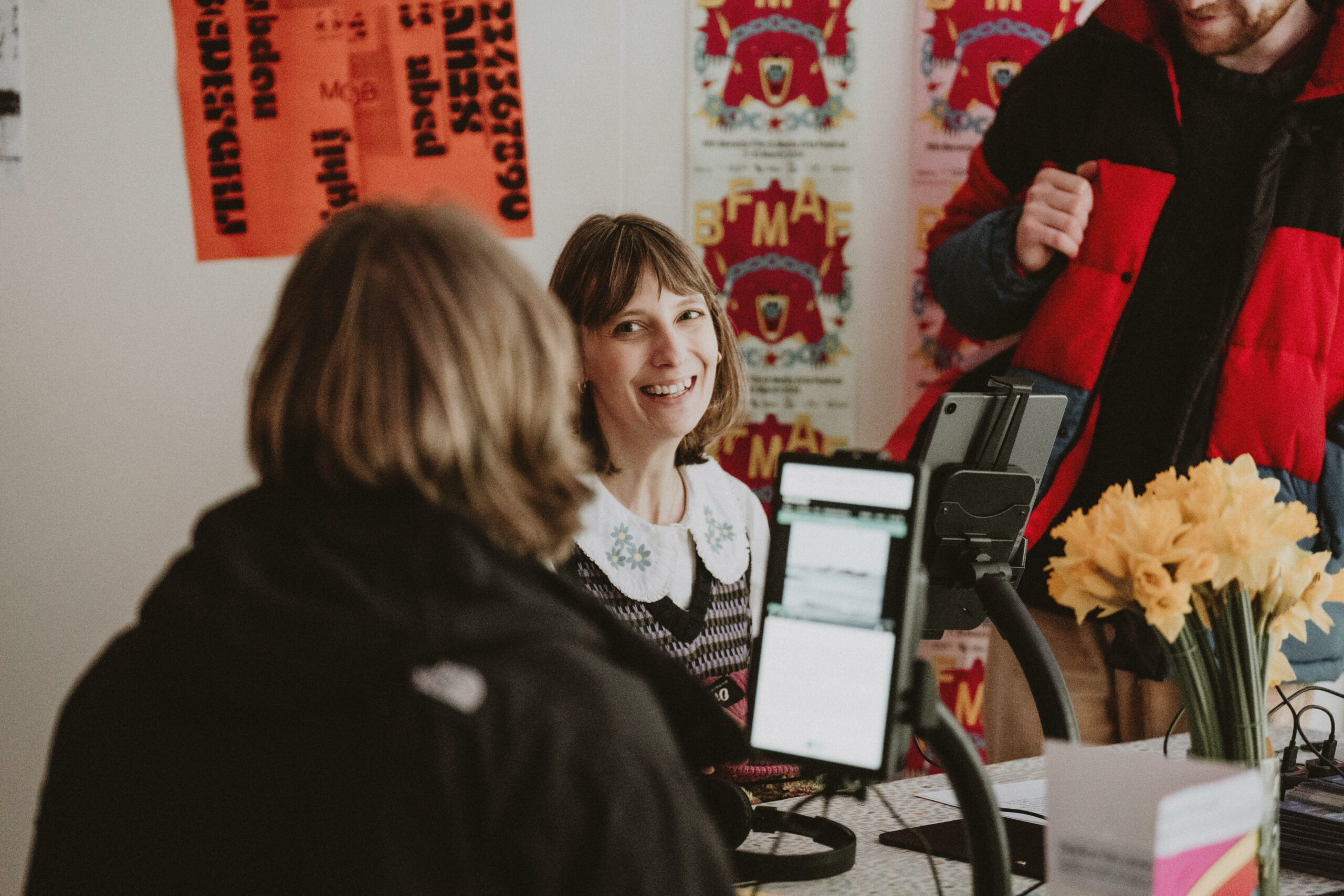 Smiling woman and the back of another persons head sat in front of a touch screen. Posters of the bright pink Berwick bear Festival poster in the background.