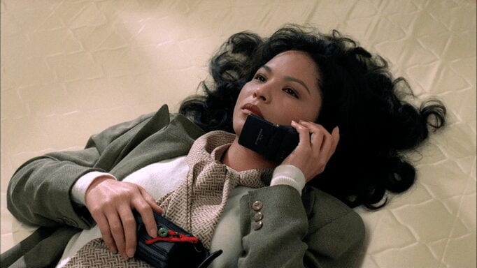 Woman lying on a light brown floor dressed in a grey suit with a 90s mobile phone to her ear