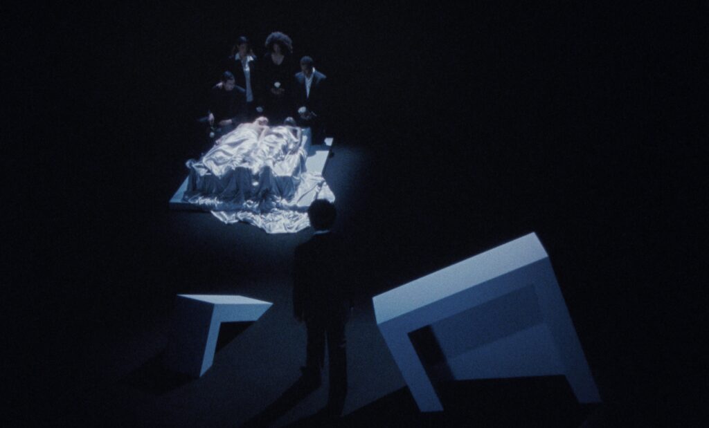 surreal image of two people laid down under a white sheet with people surrounding them in a black room