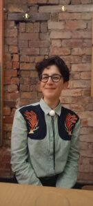 Headshot of Ghada smiling, with short black hair, glasses and wearing a light blue shirt.