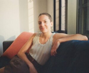 Headshot of Milda reclining on a sofa, with cropped hair and an off-white sleeveless shirt.