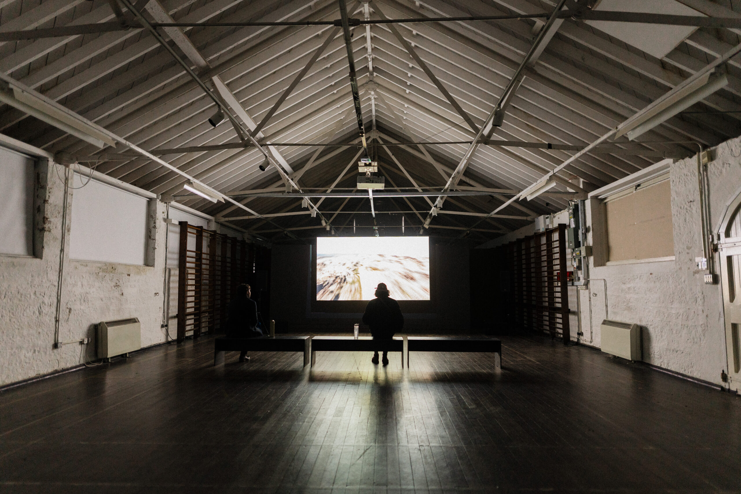 Cavernous empty warehouse space with a screen and silhouette of one person watching it.