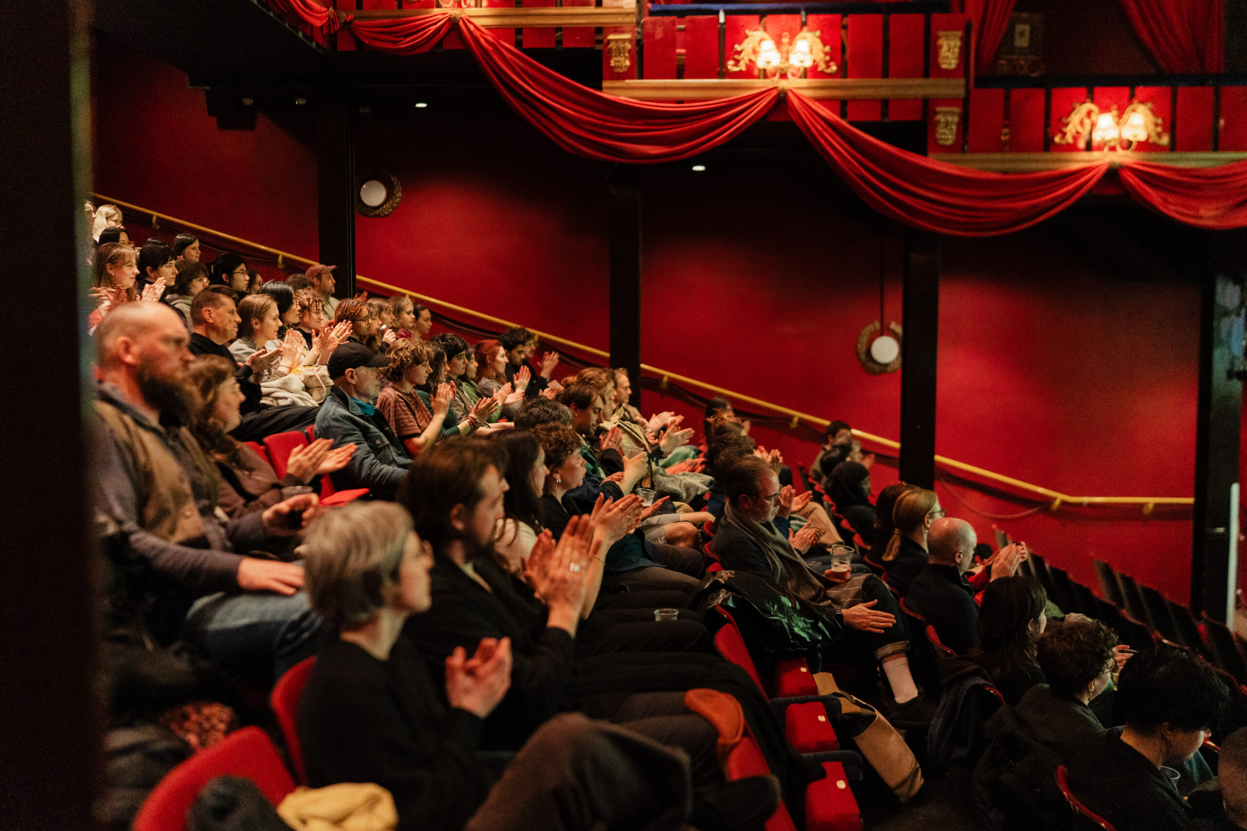 Side-view of cinema audience applauding. Deep red upholstery and walls.