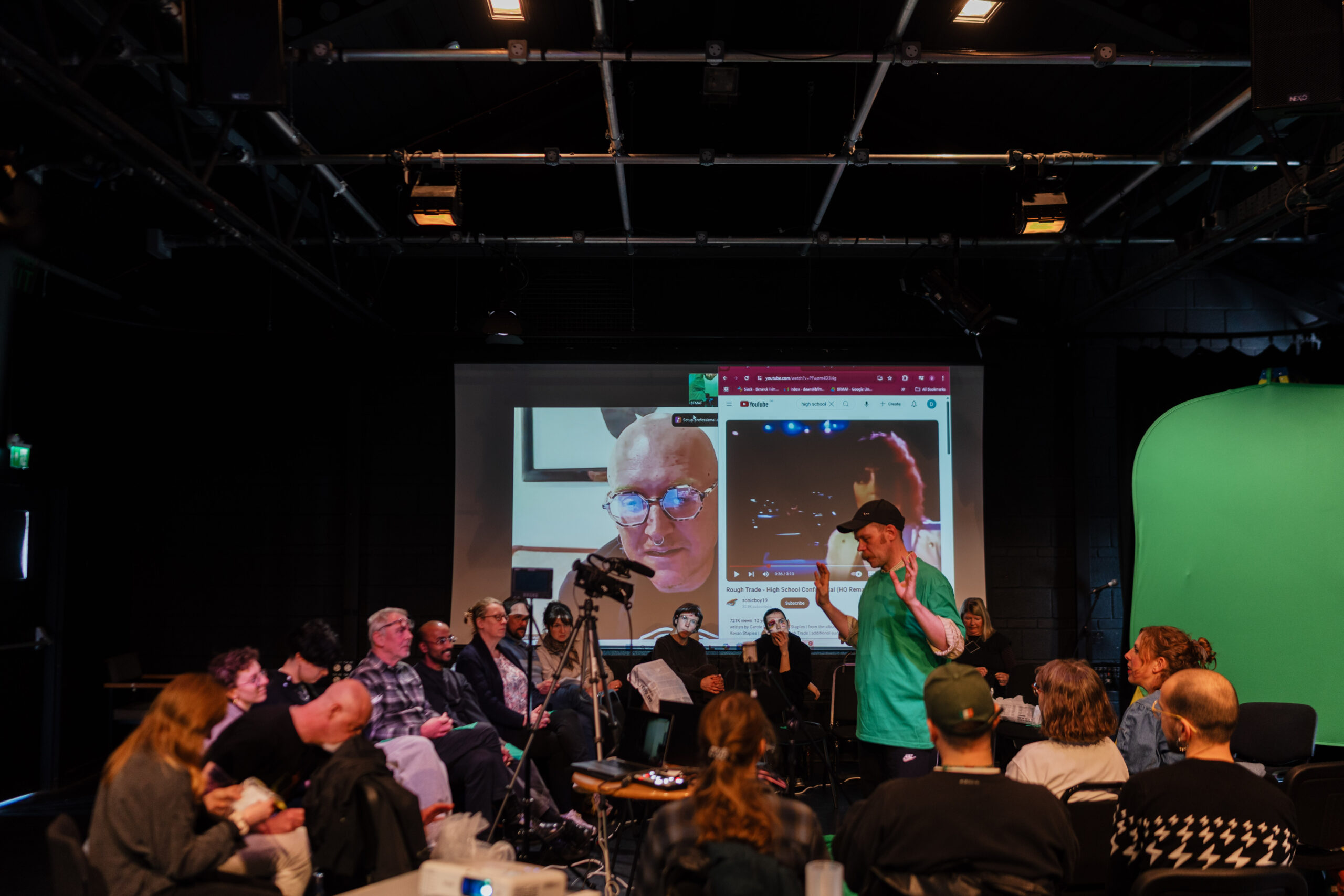 Conal McStravick in a green t-shirt leading a group of people in a workshop. Screen behind them displays a zoom call and webpage.