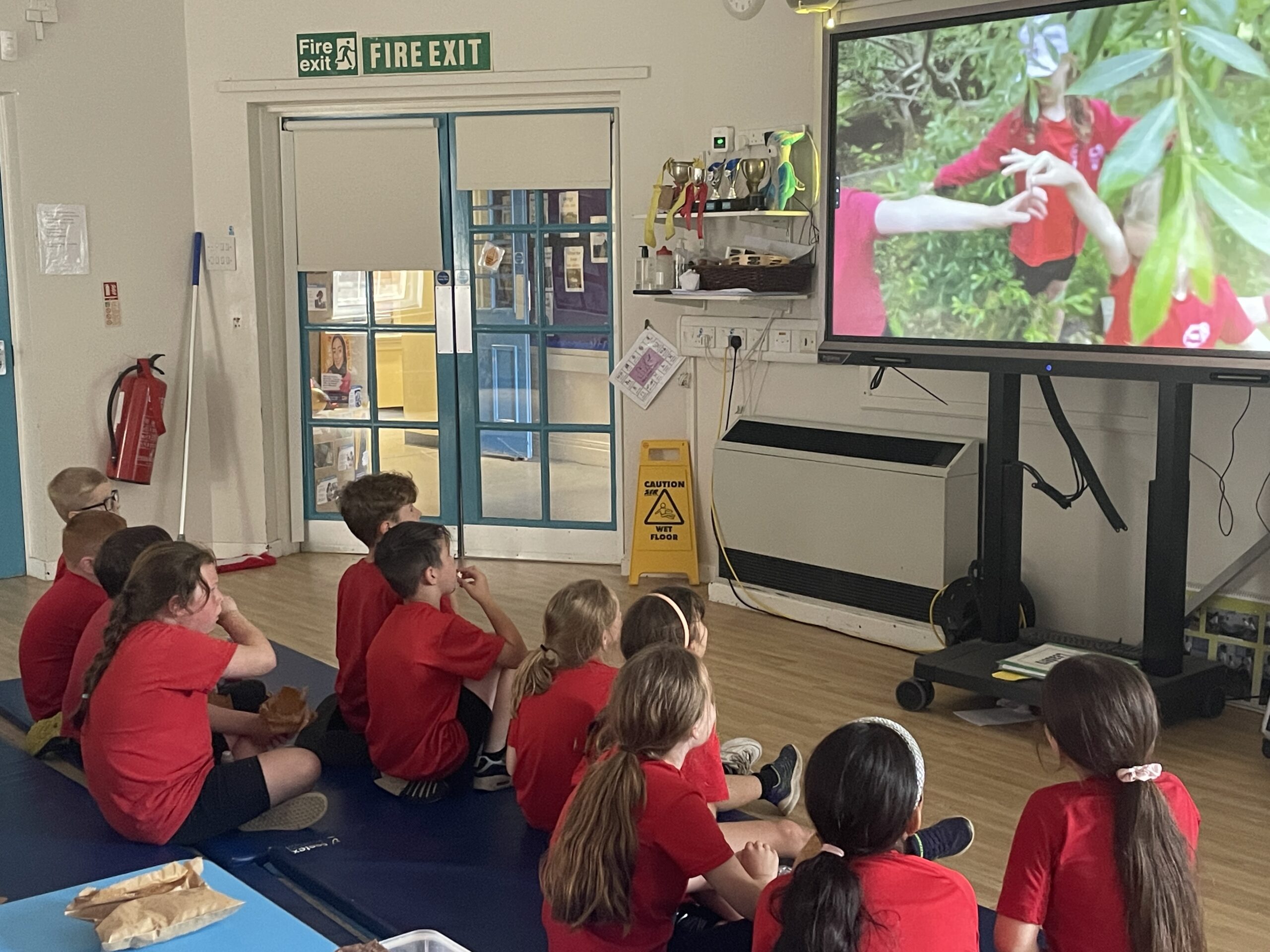 children watching a tv screen wearing red shirts and sat on the floor of a classroom