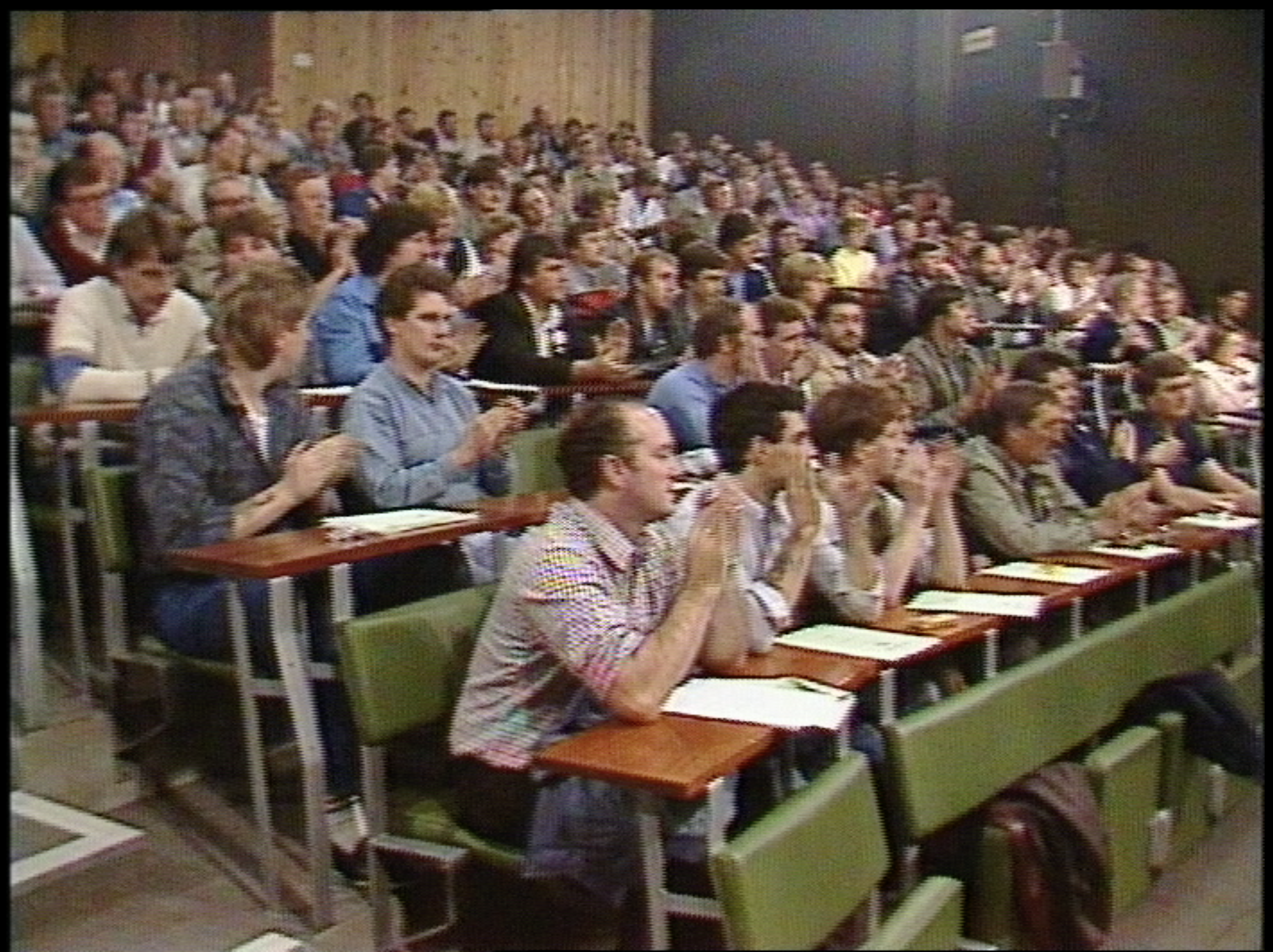 a crowd of people sat on university-style bleachers with green chairs and desks