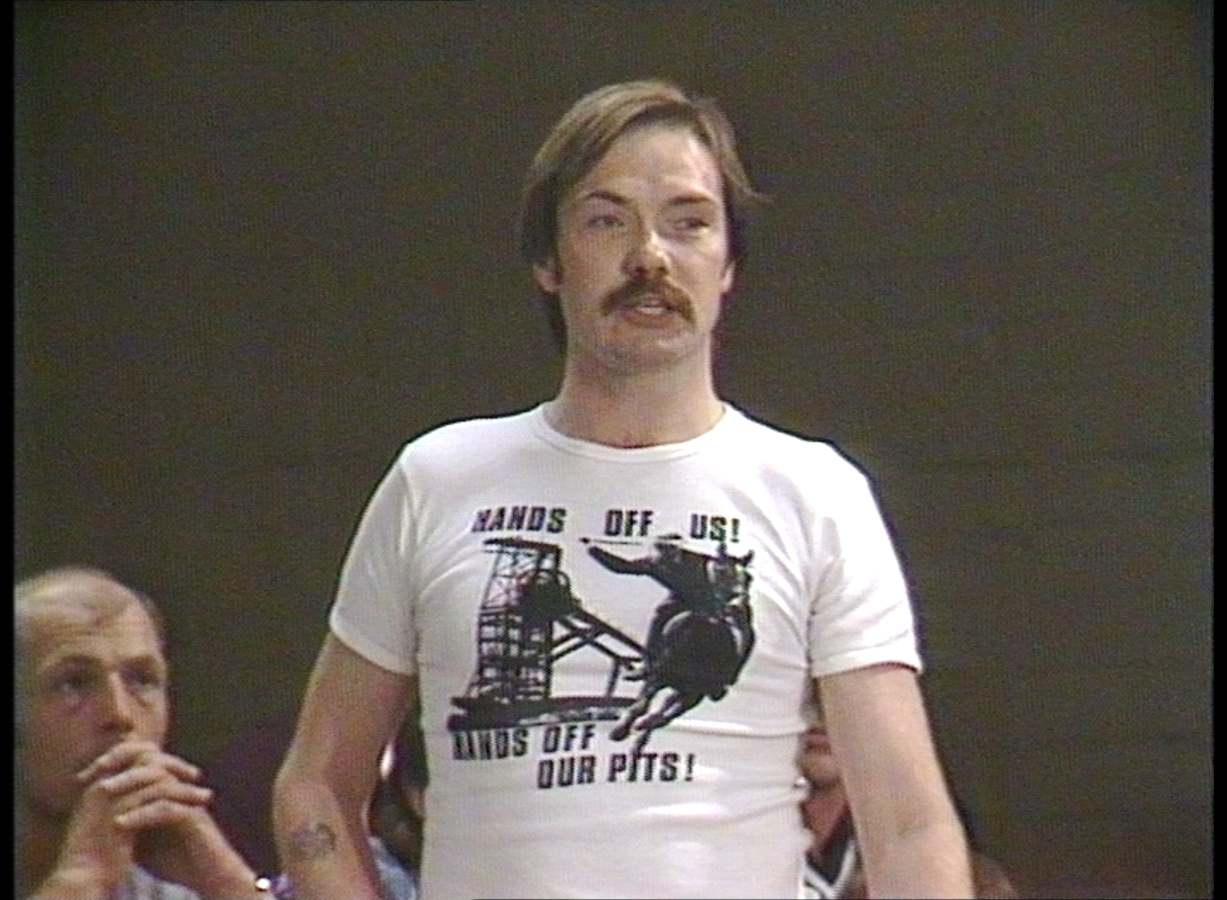 man with dark hair and moustache standing. his white t-shirt reads "Hands off us, hands off our pits!"