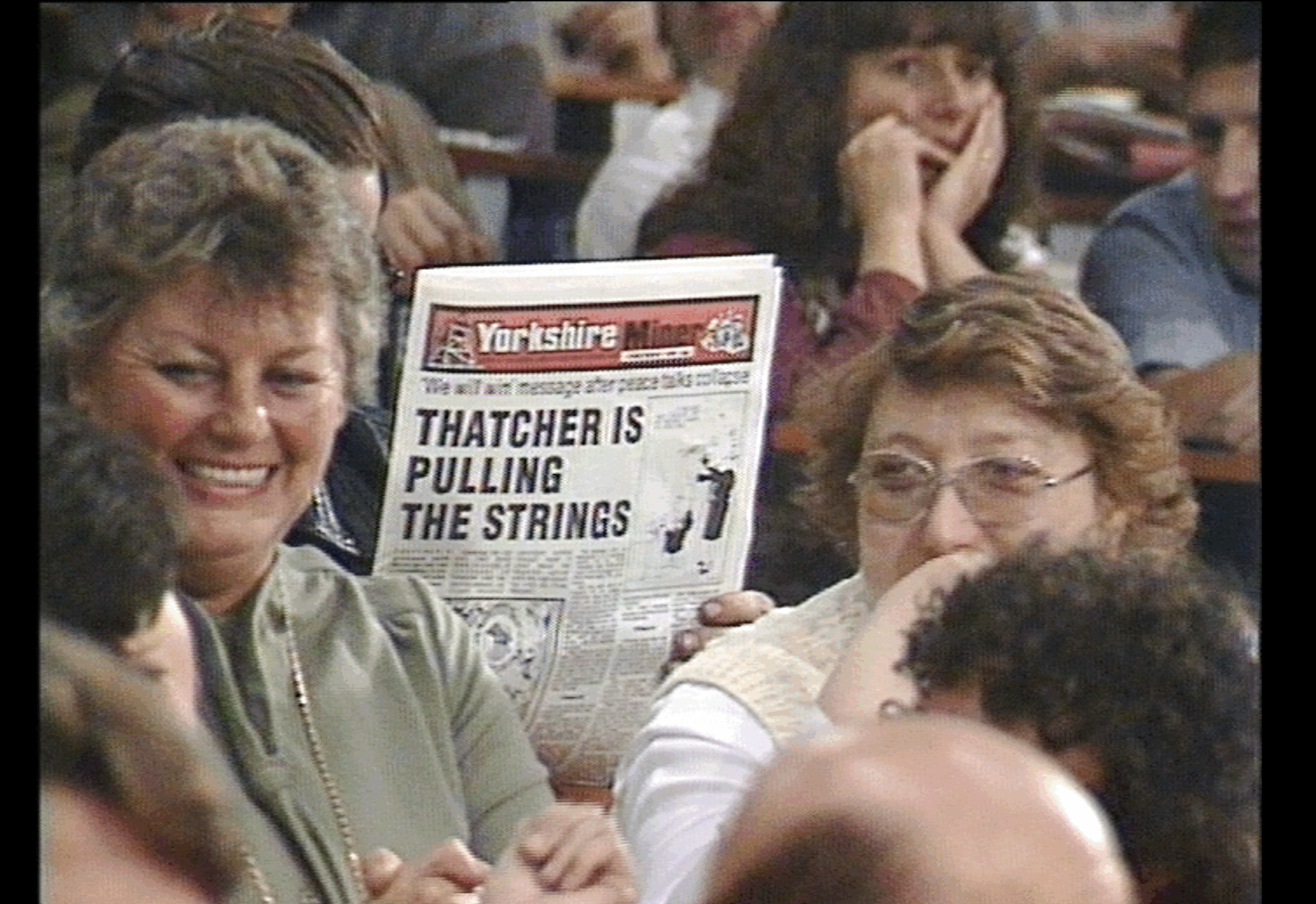Amongst a crowd, two women smiling. someone holds a paper up behind them, the headline reads "Thatcher is Pulling Strings"