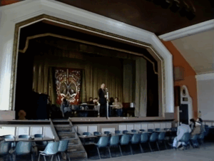 A hall with a stage and someone at the microphone