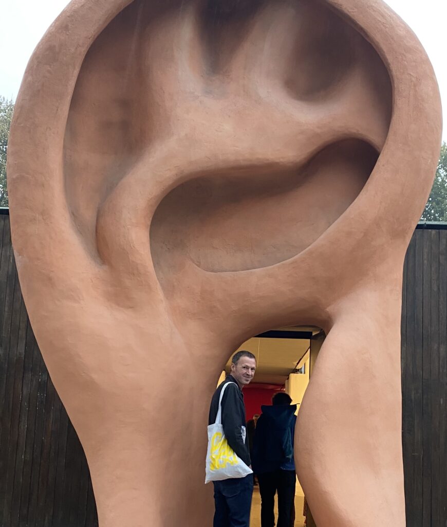 Mal standing inside the cavity of a giant ear, holding a BFMAF tote bag