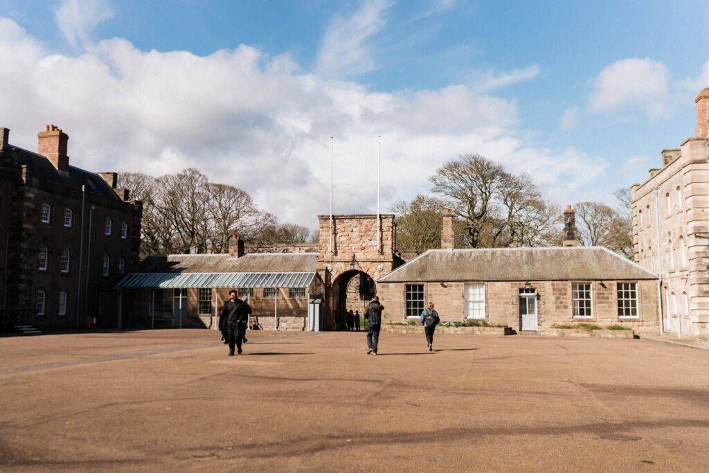 Three adults walking in a spacious historic courtyard with stone buildings and leafless trees.