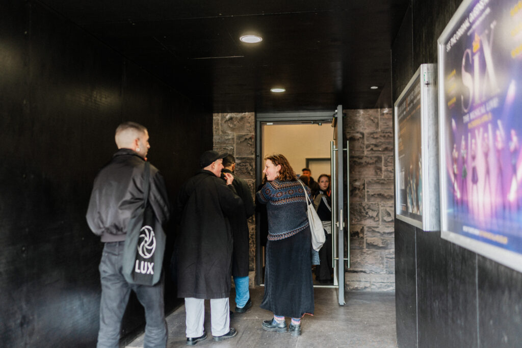 A group of adults walking through a dark hallway into a theater entrance. People chatting and waiting to enter The Maltings' Cinema at the Barracks. Audience members arriving at a cultural event.