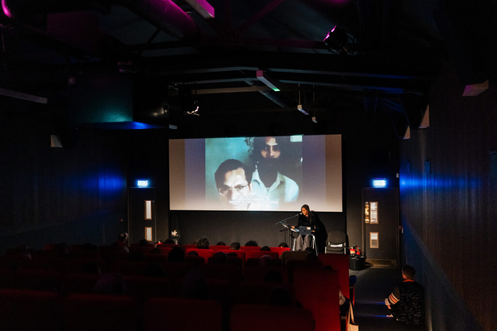 Abiba Coulibaly speaking at microphone during "Black and Arab Encounters on Screen" in dark theater. Audience seated in red chairs watching movie with subtitles. Public event with presentation and film viewing.
