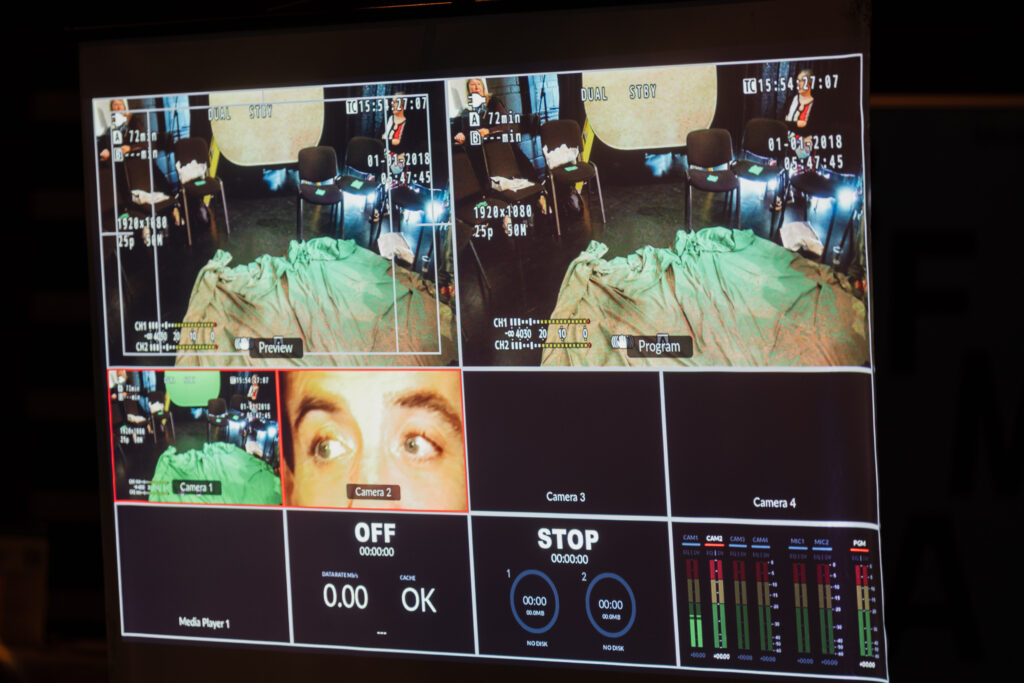 Video production monitor displaying multiple camera feeds including a close-up of a man's eyes and an empty chair setup. Studio recording environment with technical overlays and audio levels.