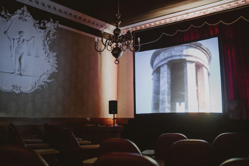 darkened cinema space inside the town hall with emblem on the left hand wall, chandelier haning from the ceiling and image of a building on screen