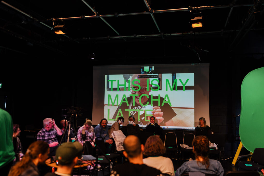 Group of adults attending a film festival panel discussion in a dark room with a large screen showing a video call. People listening, seated and engaged in conversation. Film event gathering.