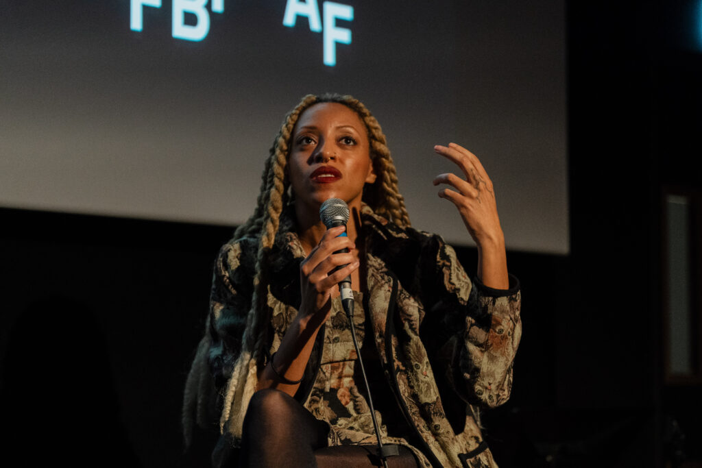 Woman with long blonde braids holding microphone and speaking during film festival panel. Female speaker sharing ideas at event. Public speaking and cultural discussion.