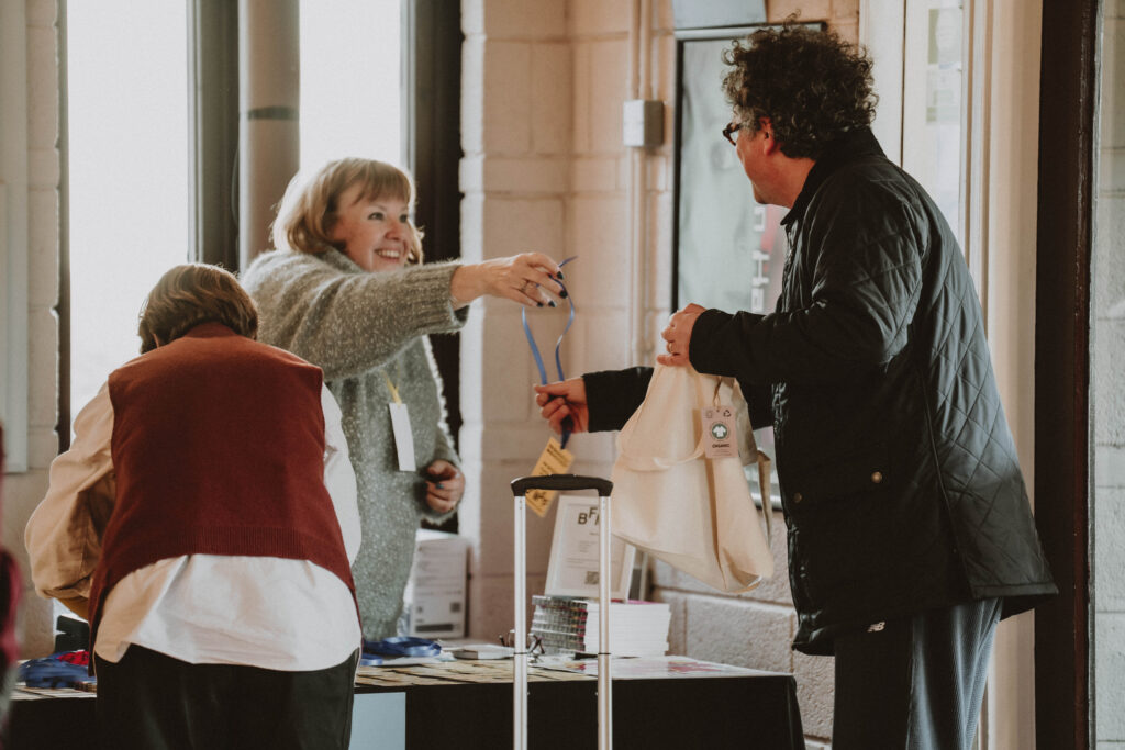 Smiling woman handing a blue lanyard badge to a man at a film festival registration desk. Event staff assisting guests with passes and tote bags. Festival check-in and welcoming scene.