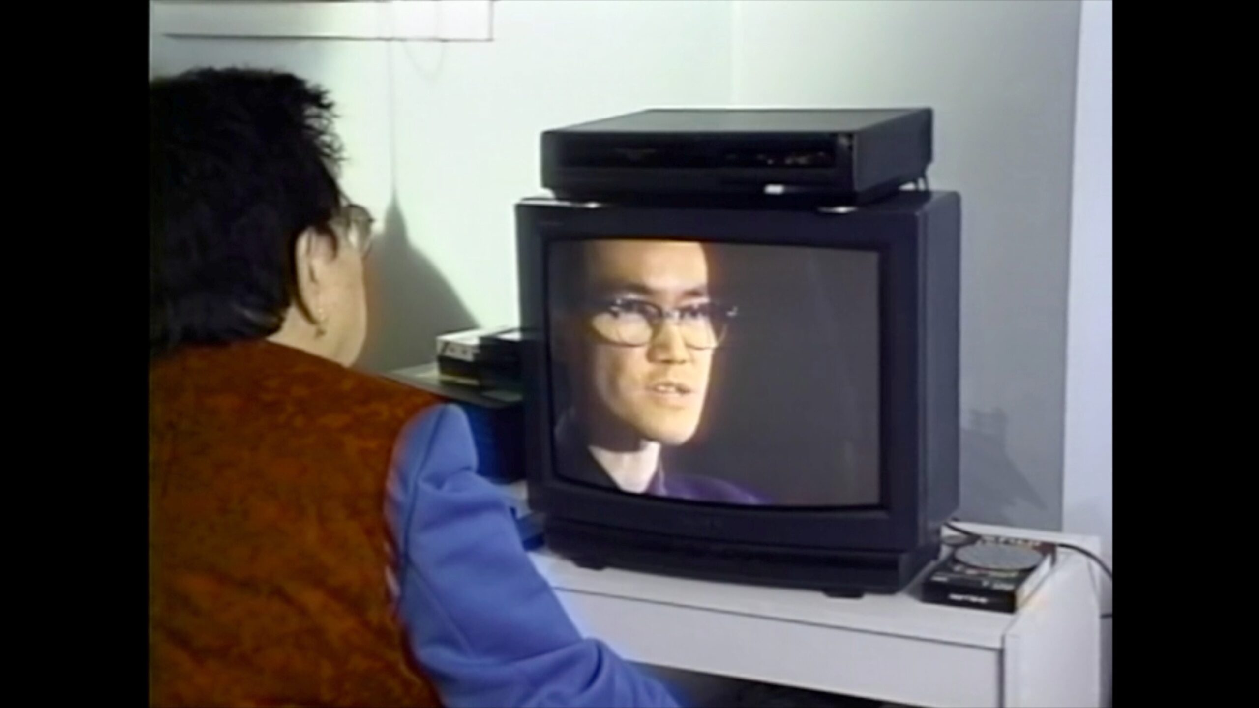Adult watching a man speaking on an old CRT television screen. Person seated at a desk with VHS player and tapes nearby. Watching vintage media in a home setting.