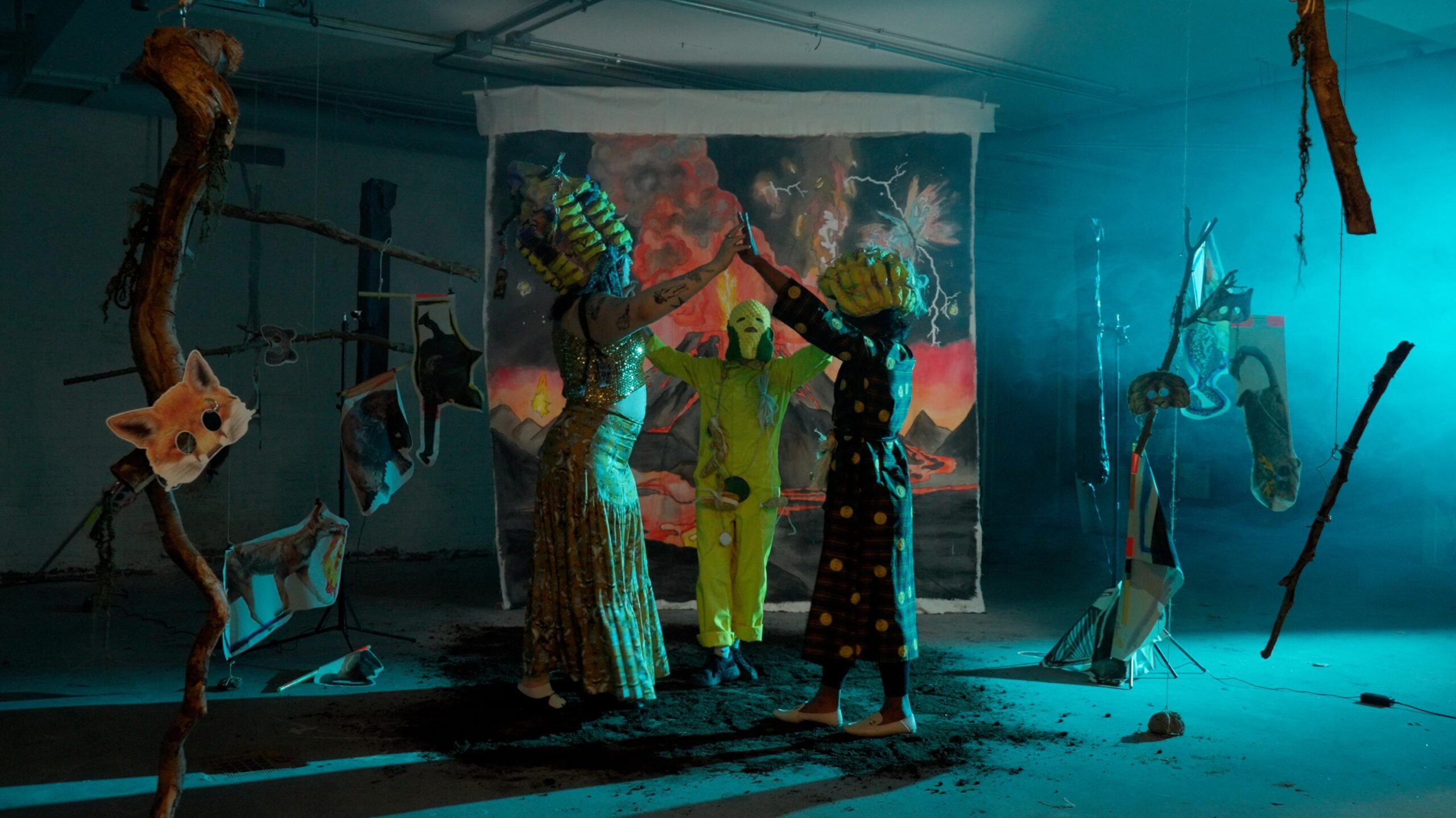 Three adults wearing colorful masks and costumes perform in a dimly lit art installation with hanging objects and a painted backdrop. Creative theatrical scene with surreal elements.