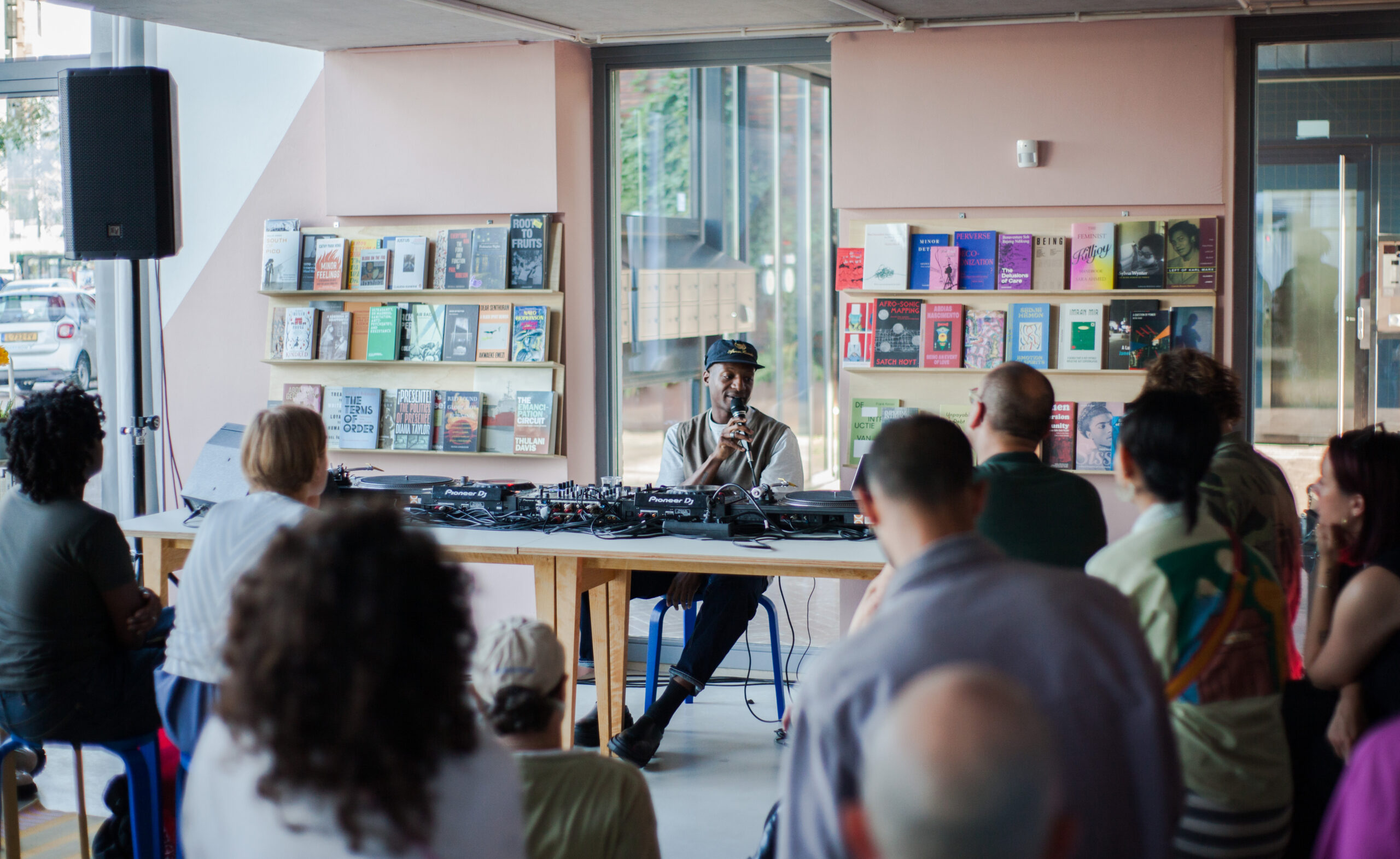 Ashley Holmes sitting in a room with books and zines behind him and a table with decks in front, addressing a crowd of people