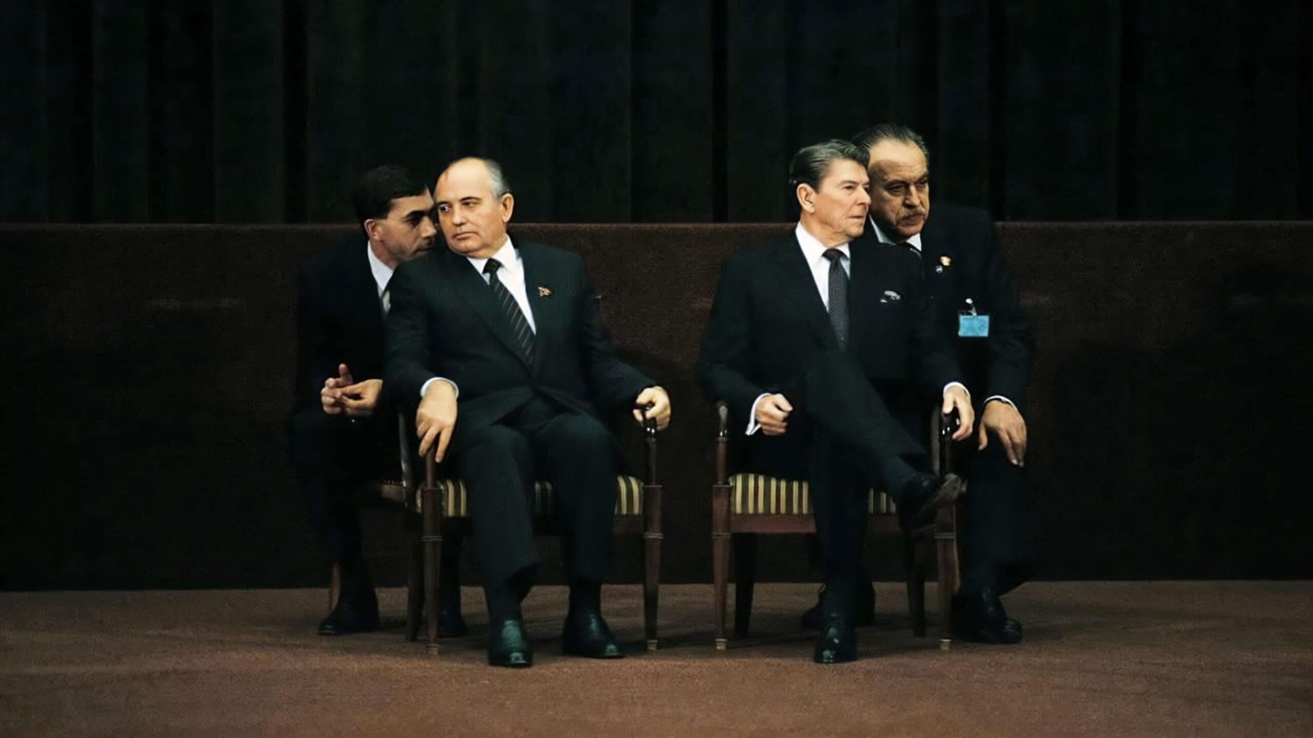 film still displaying four men in dark formal suits seated on stage chairs, two men whispering to seated leaders. Political meeting scene with serious expressions and intense body language.