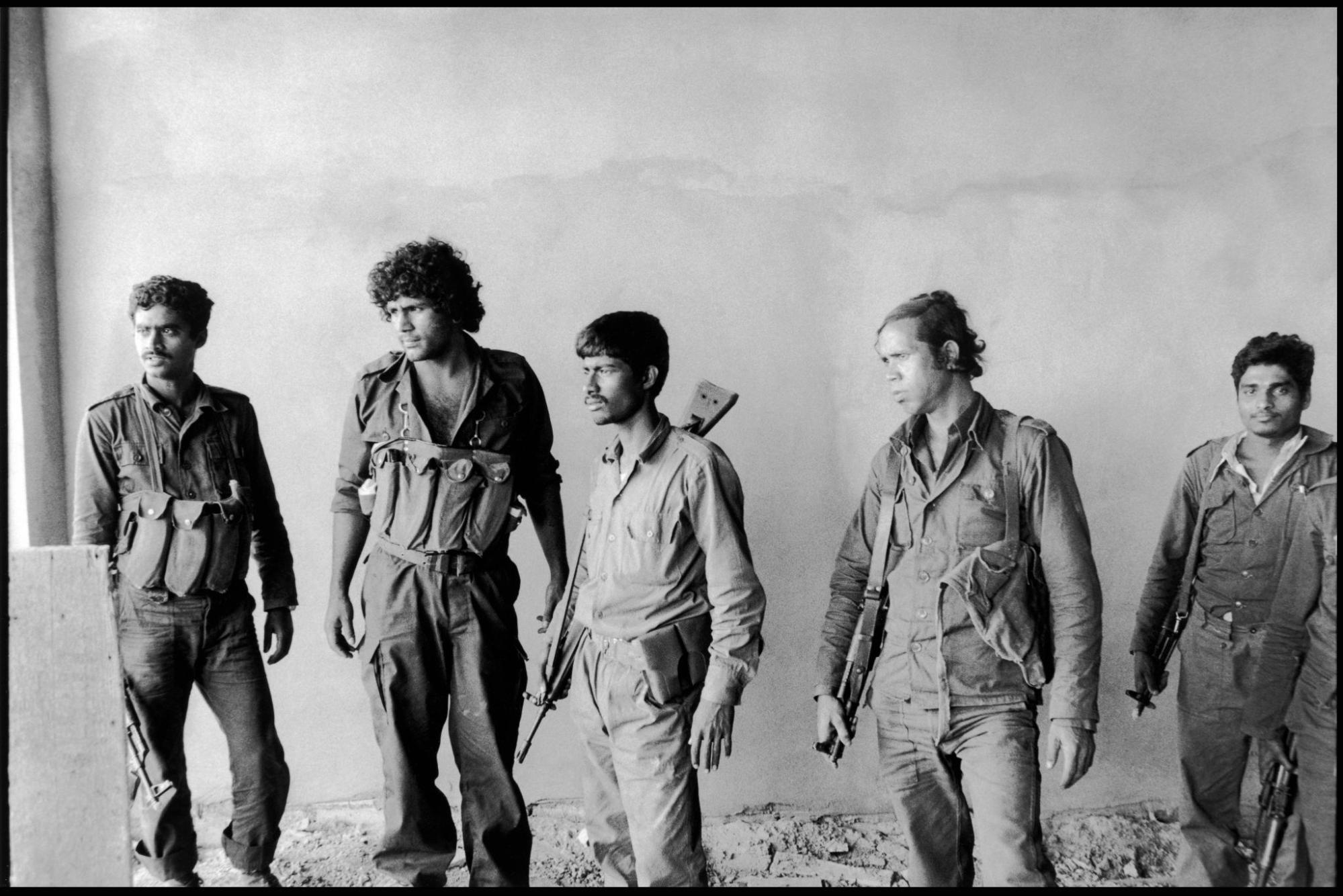 Five armed adult men in military-style clothing stand inside a bare room. They appear alert and focused, possibly preparing for or recovering from an operation. Soldiers with rifles and tactical gear.